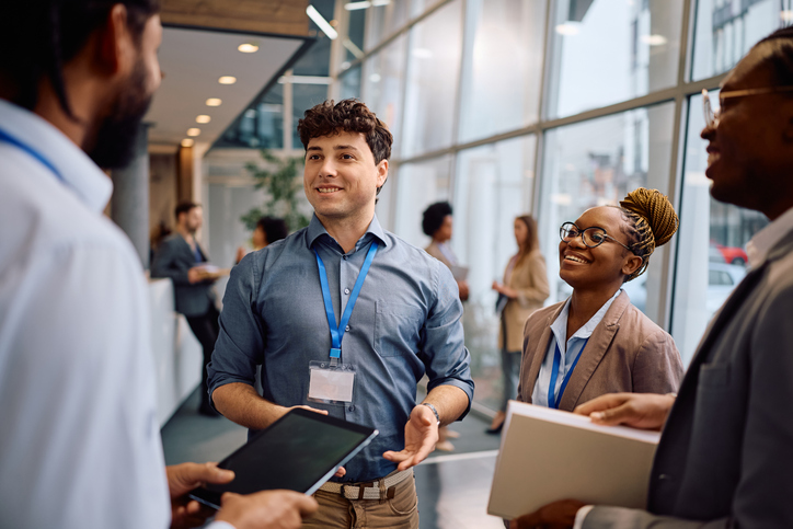 Diverse professionals in conference setting, engaging in conversation, holding notebooks and devices, with name tags visible.