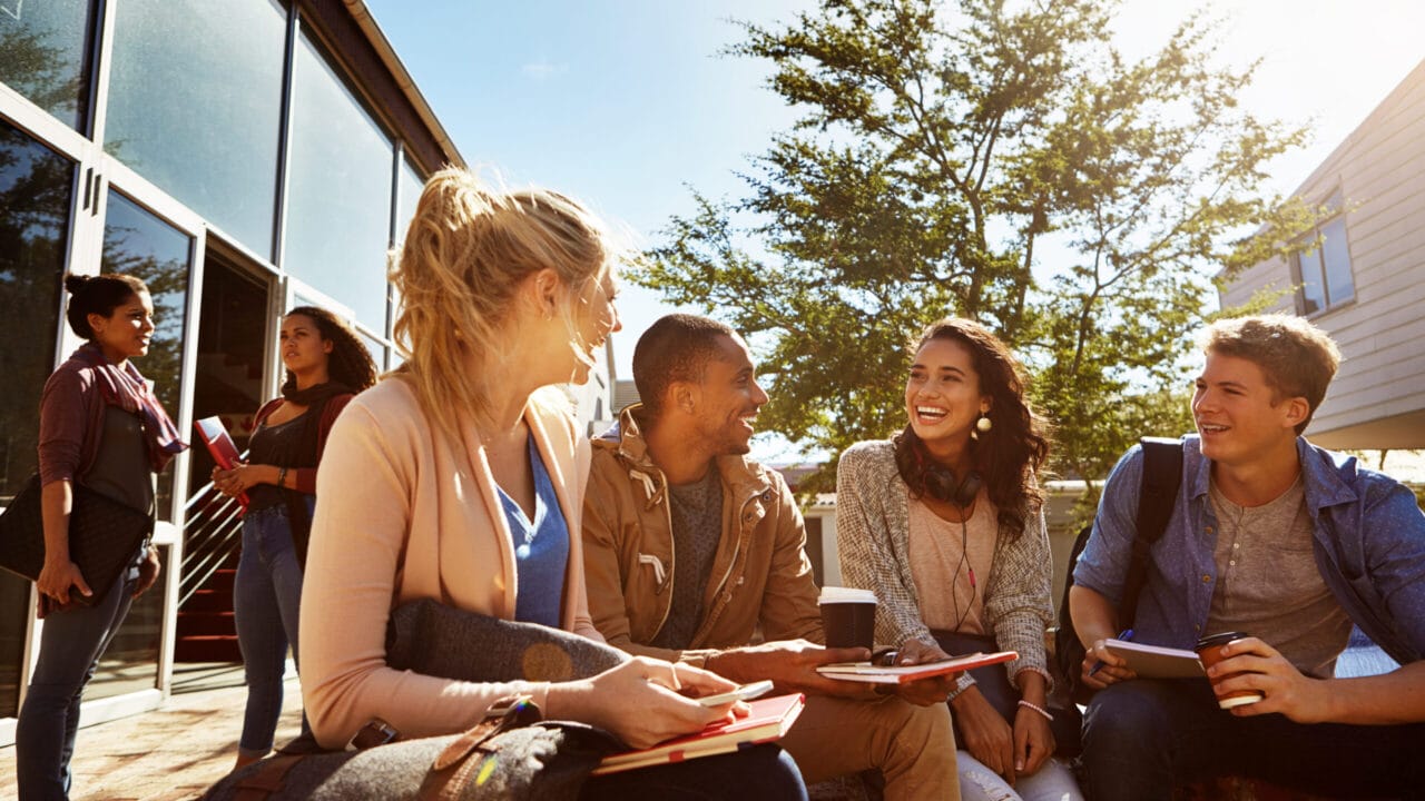 Group of young adults sitting outdoors, chatting and smiling, with books and coffee under sunny skies near a modern building.