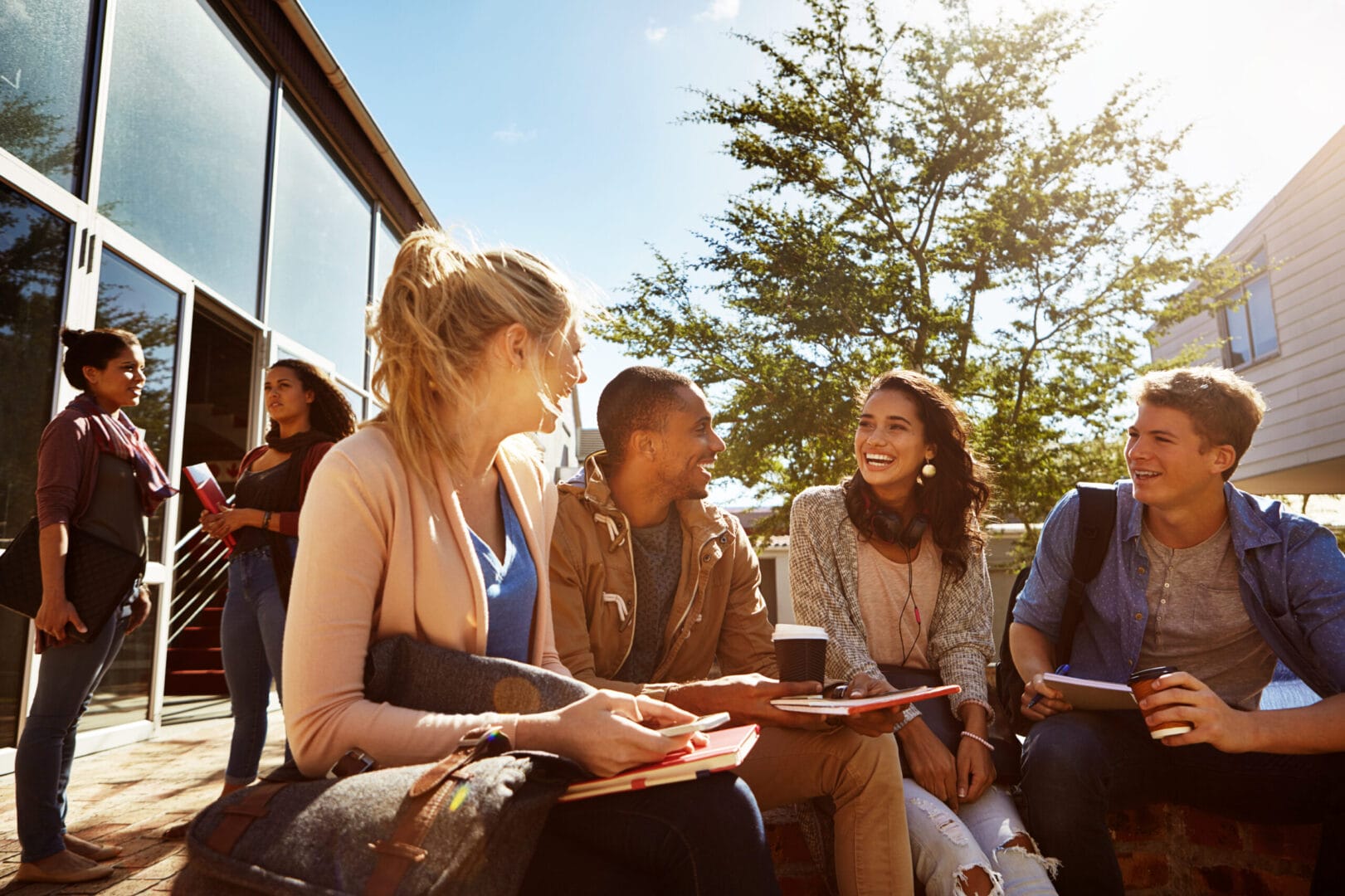 Group of young adults sitting outdoors, chatting and smiling, with books and coffee under sunny skies near a modern building.
