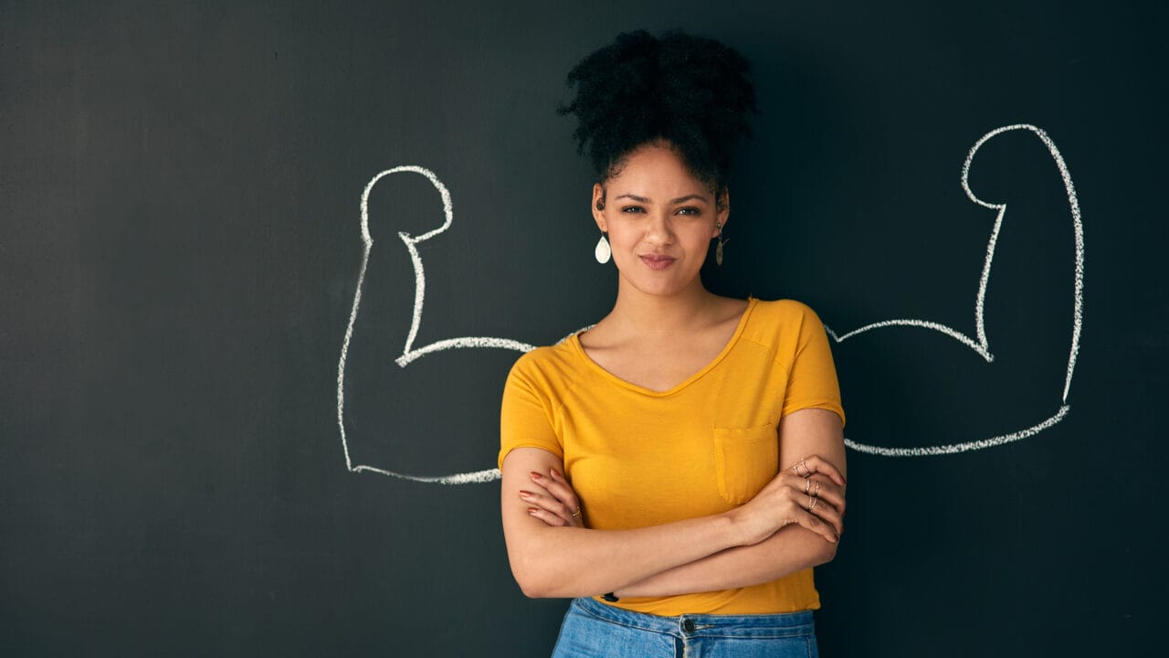 Confident woman with arms crossed stands in front of blackboard with chalk-drawn biceps, symbolizing inner strength.