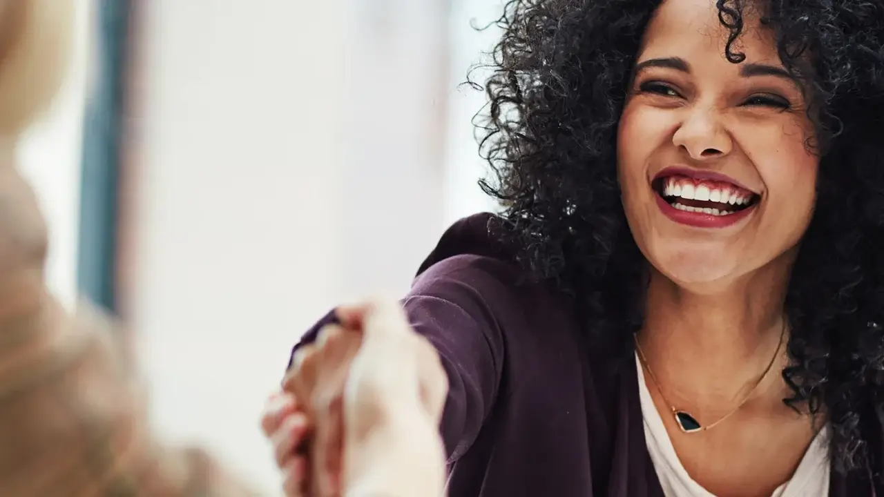 Smiling person with curly hair shaking hands, wearing a dark jacket and white shirt, expressing warmth and friendliness.
