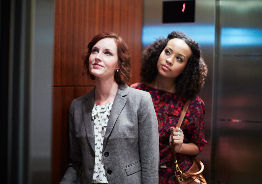 Two women in an elevator, one in a gray blazer and the other in a red floral dress, standing near a wooden panel.