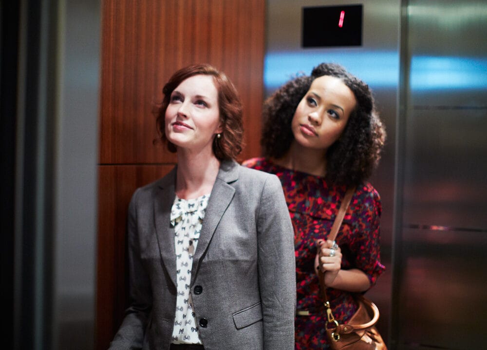 Two women in an elevator, one in a gray blazer and the other in a red floral dress, standing near a wooden panel.