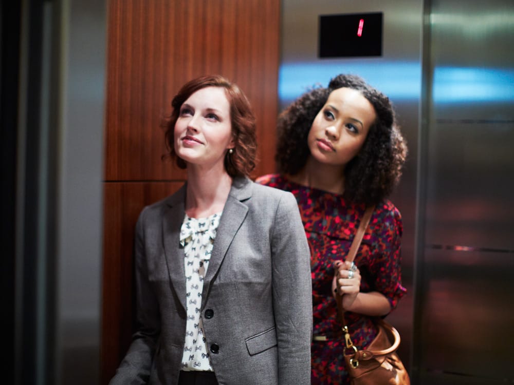 Two women in an elevator, one in a gray blazer and the other in a red dress, looking up towards the floor indicator.