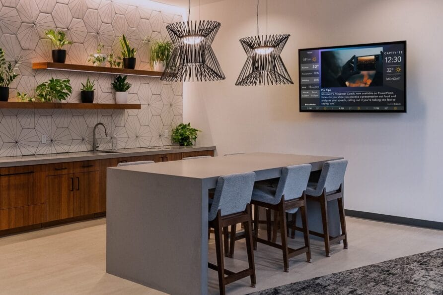 Modern kitchen with a central island, hanging lights, wall-mounted TV, and shelves with potted plants.