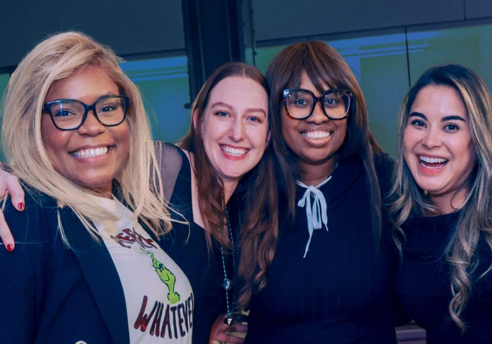 Four smiling people posing together indoors, wearing casual and professional attire.