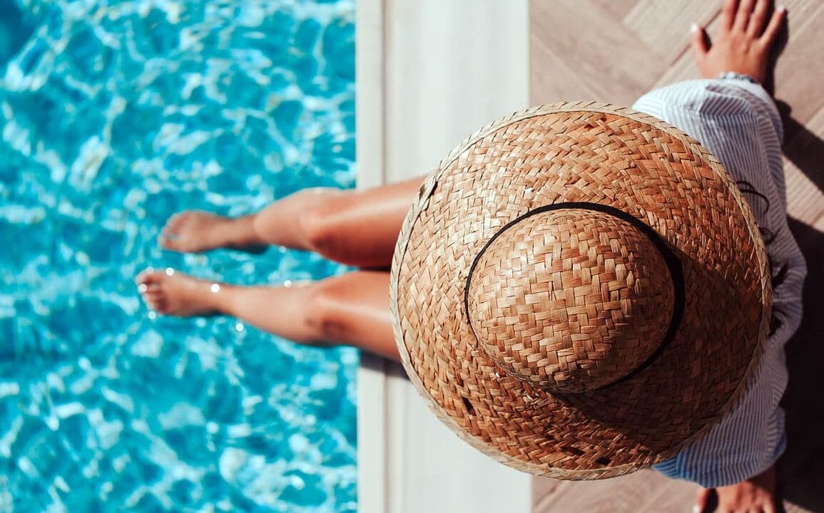 Person relaxing by a poolside, wearing a straw hat and striped shirt, legs dipped in clear blue water.
