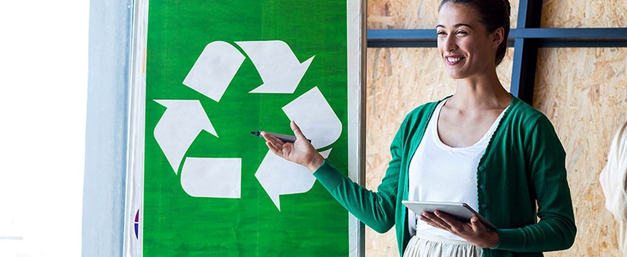 Woman presenting recycling initiative to colleagues with a green recycling symbol poster in the background.