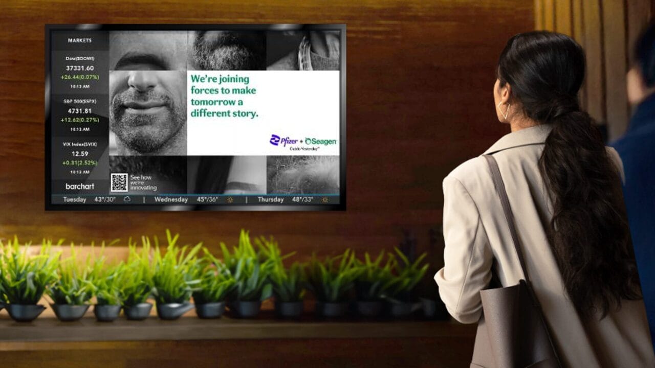 Woman observing a digital wall display with stock market info and an ad for a corporate collaboration among potted plants.