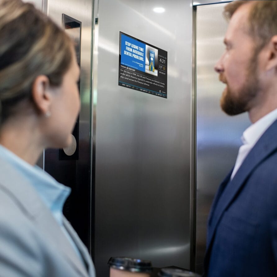 Two professionals in an elevator looking at a digital screen displaying information.
