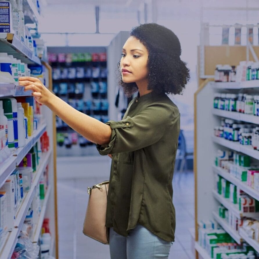 Woman browsing products on a pharmacy shelf, wearing a green shirt and carrying a beige bag.