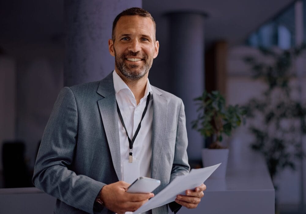 Smiling man in a suit holding papers and a smartphone in an office setting.