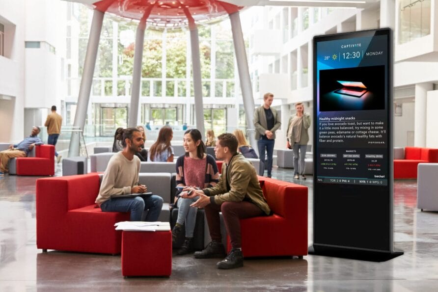 People sitting on red couches in a modern lobby with a large digital screen displaying information.
