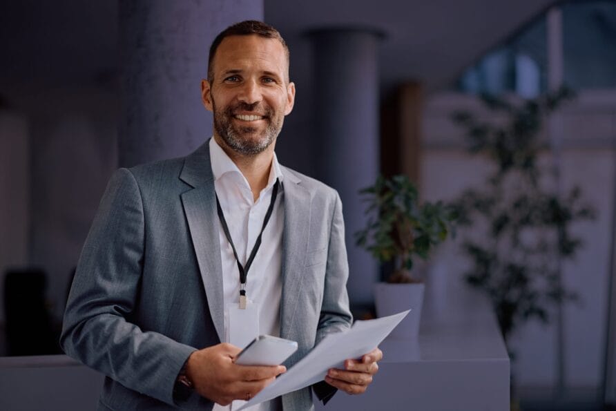 Smiling man in a gray suit holding papers and a phone, standing indoors with a potted plant in the background.