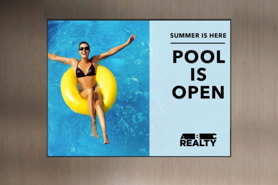 Woman in swimsuit relaxing on yellow float in pool beside sign reading "Pool is Open" from a realty company.