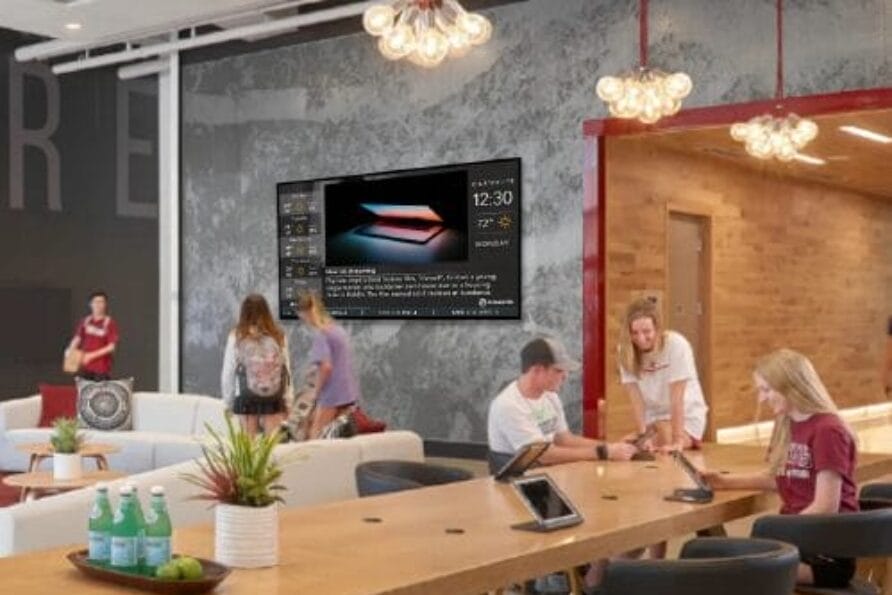 Modern office space with people working at a long table; wall-mounted screen displaying a presentation in the background.