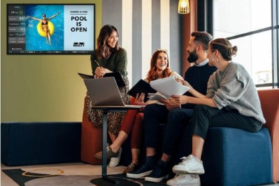 Group of colleagues sitting and talking with documents in a modern office space, poster on wall in background.