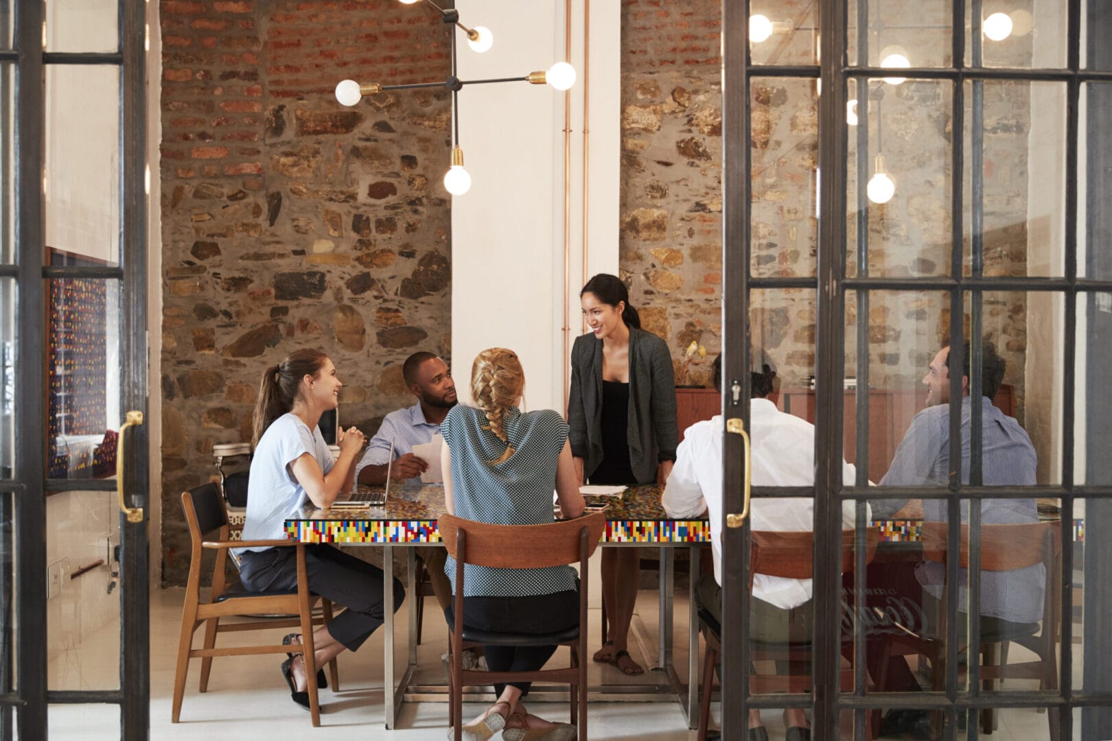 Team meeting in modern office with brick walls and glass doors, six people seated at a table discussing plans.