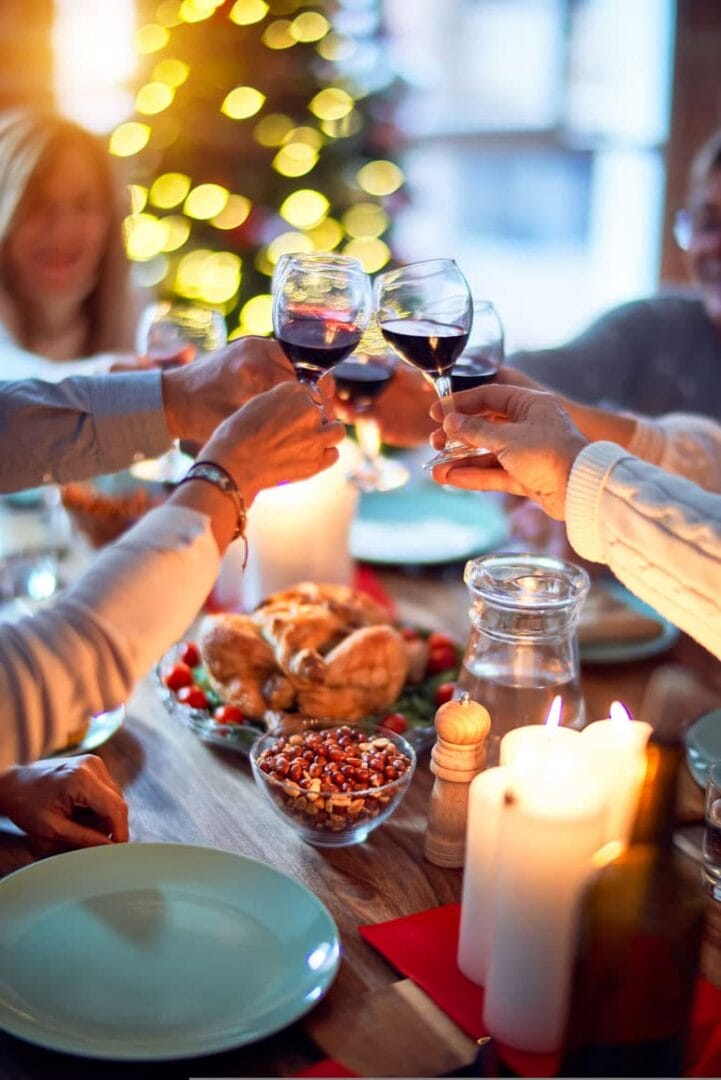 Friends toasting with wine at a festive holiday dinner table, surrounded by candles and a lit Christmas tree.