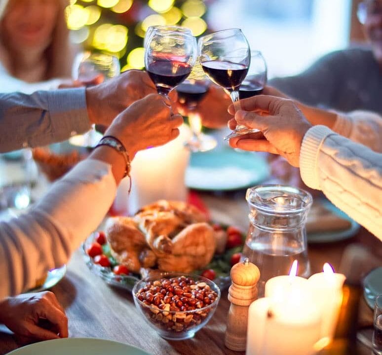 Friends toasting with wine at a festive holiday dinner table, surrounded by candles and a lit Christmas tree.
