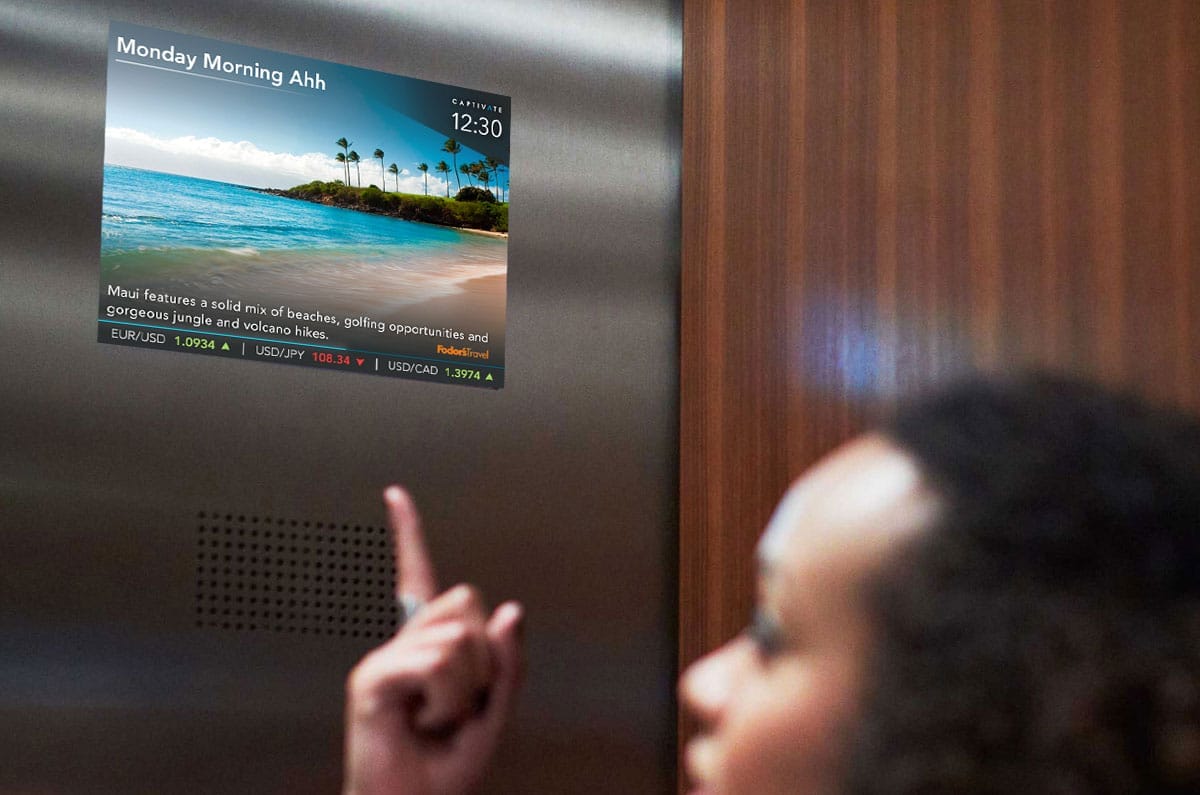 Person in elevator pointing at screen displaying scenic beach image and financial data.