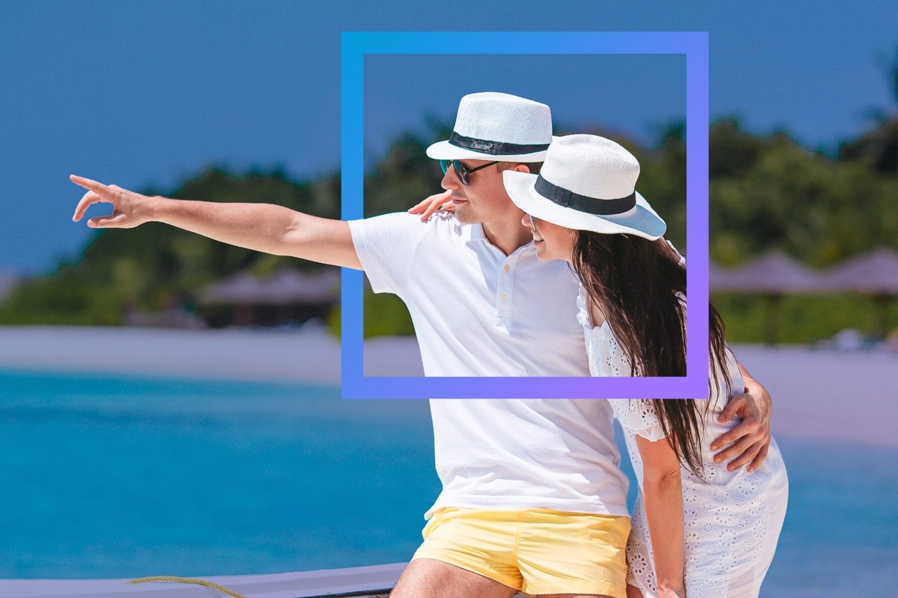 Couple in matching hats embracing on a beach, with one person pointing towards the distance, clear blue sky and water.