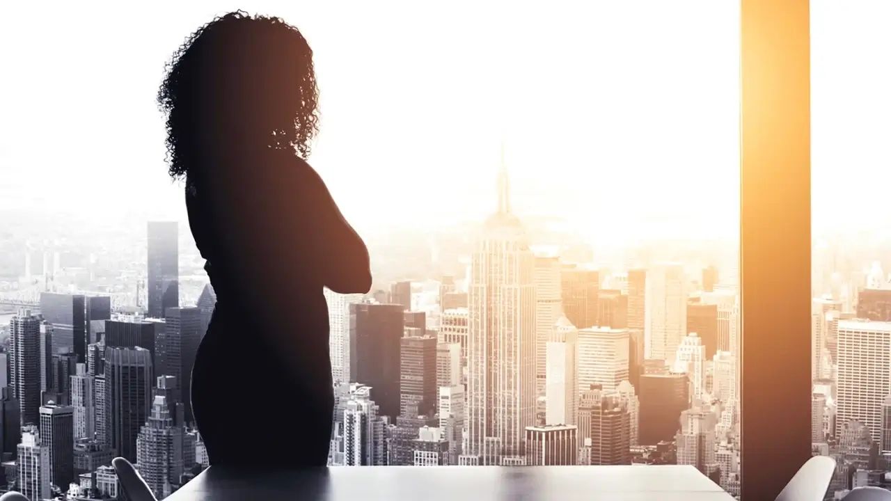 Silhouette of a person standing in front of a window overlooking a city skyline with the sun setting