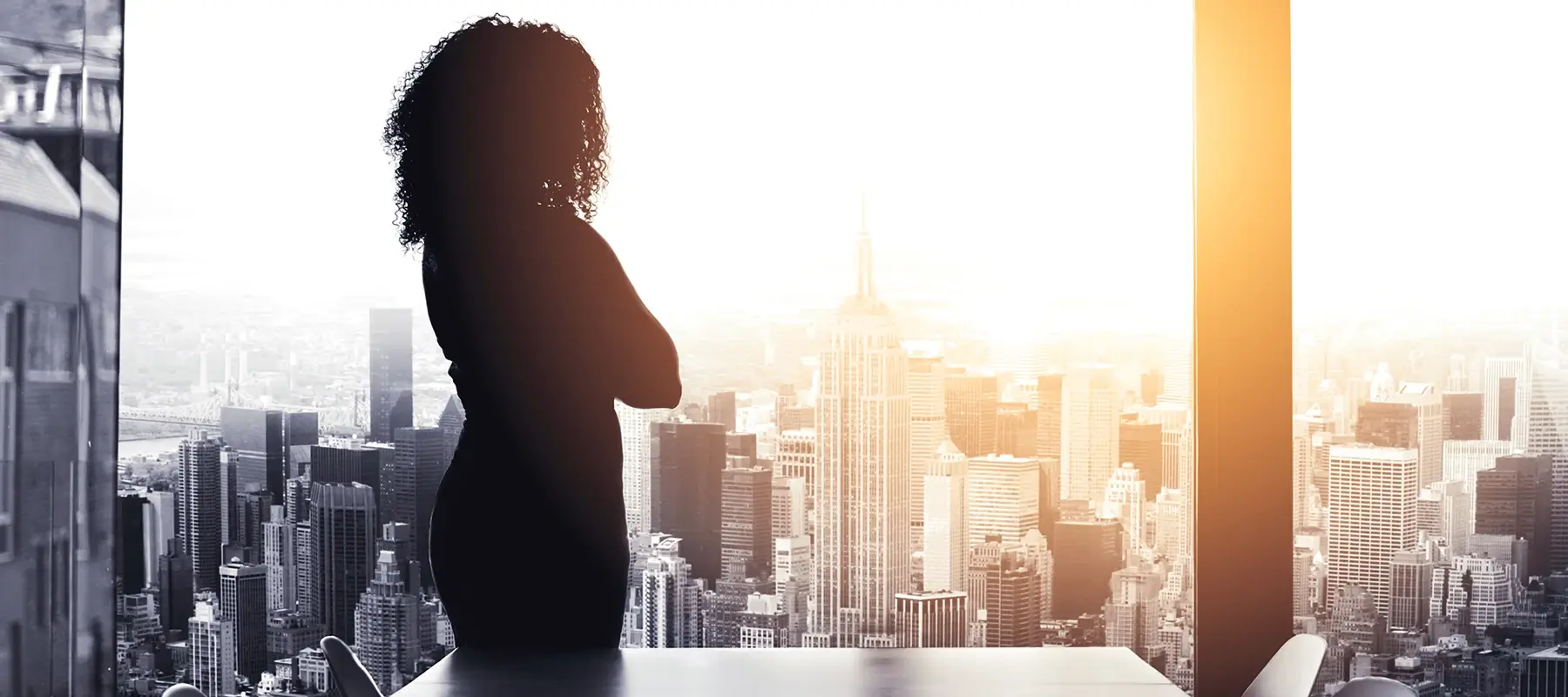 Silhouette of a person standing in front of a window overlooking a city skyline with the sun setting