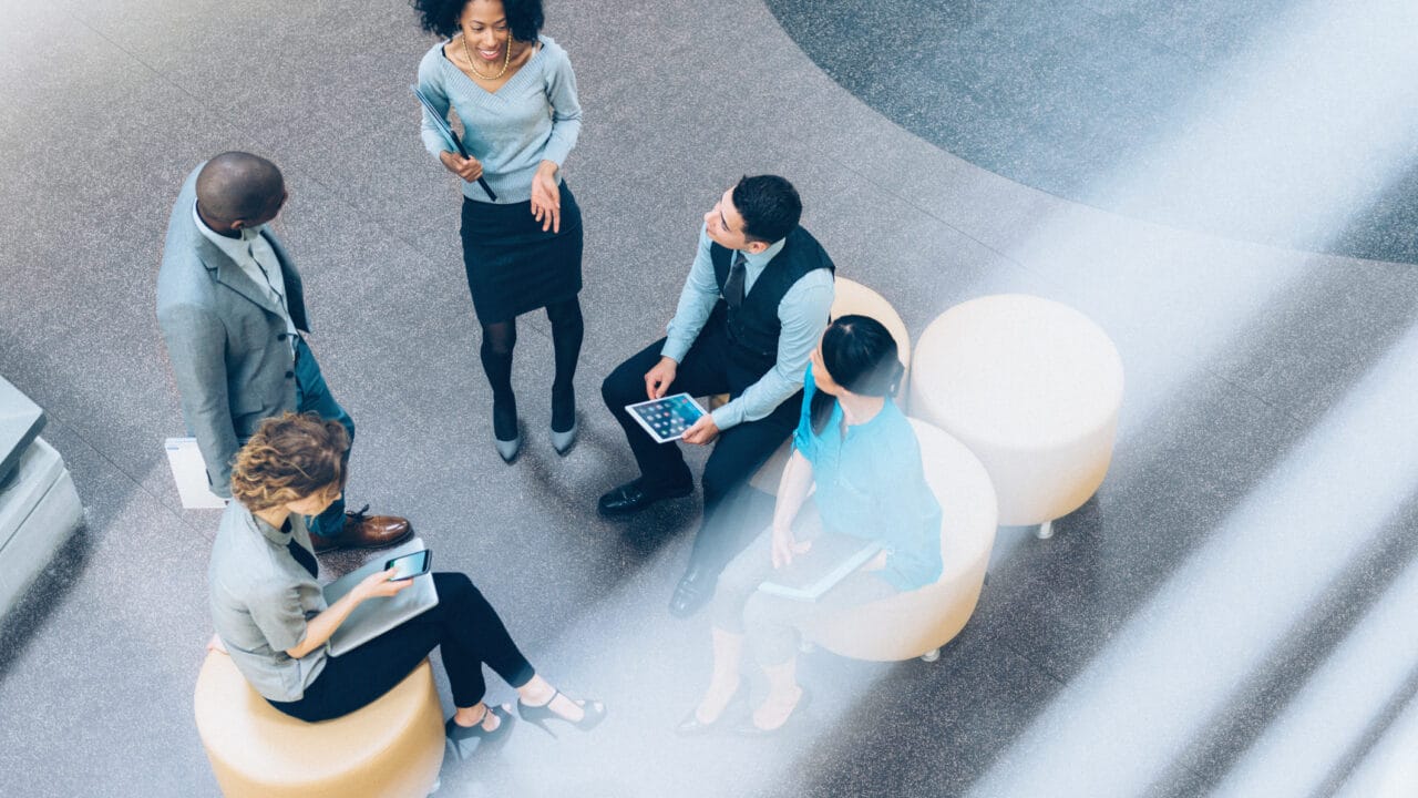 Group of five professionals having a meeting seated on round chairs, seen from above, with one person standing and speaking.