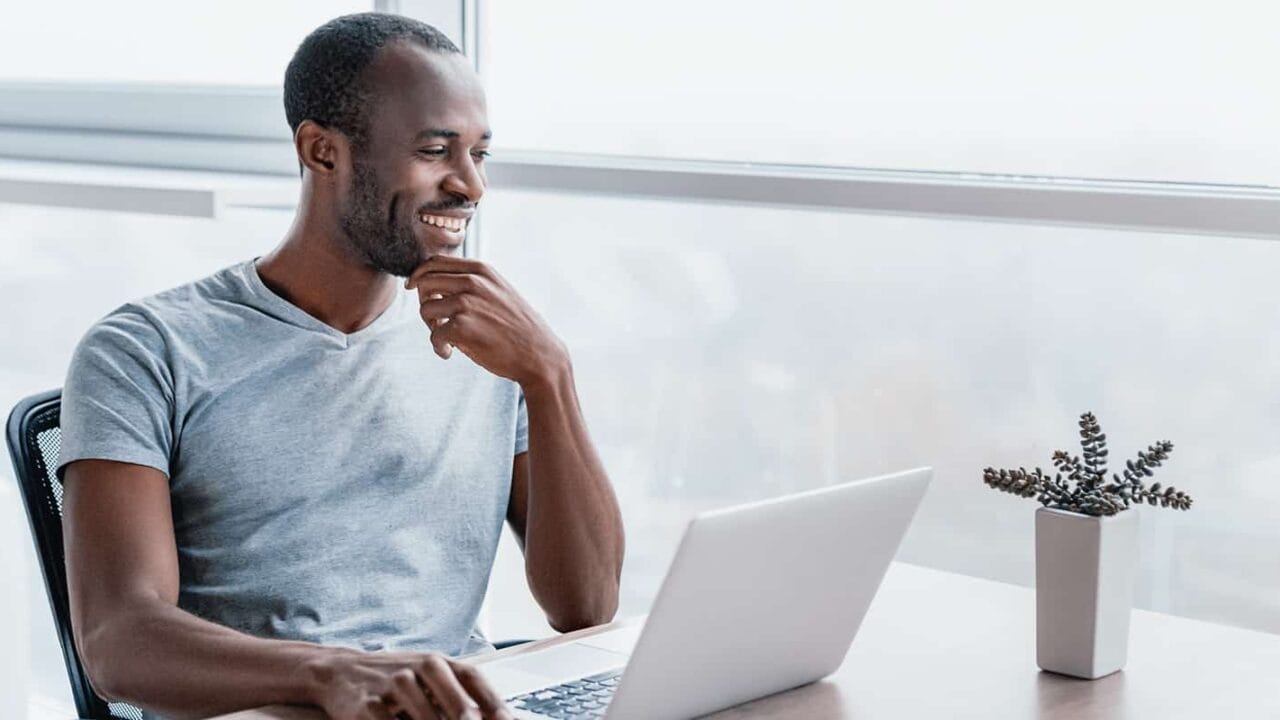 Man smiling while working on a laptop at a desk near a window with a small plant, wearing a gray t-shirt.