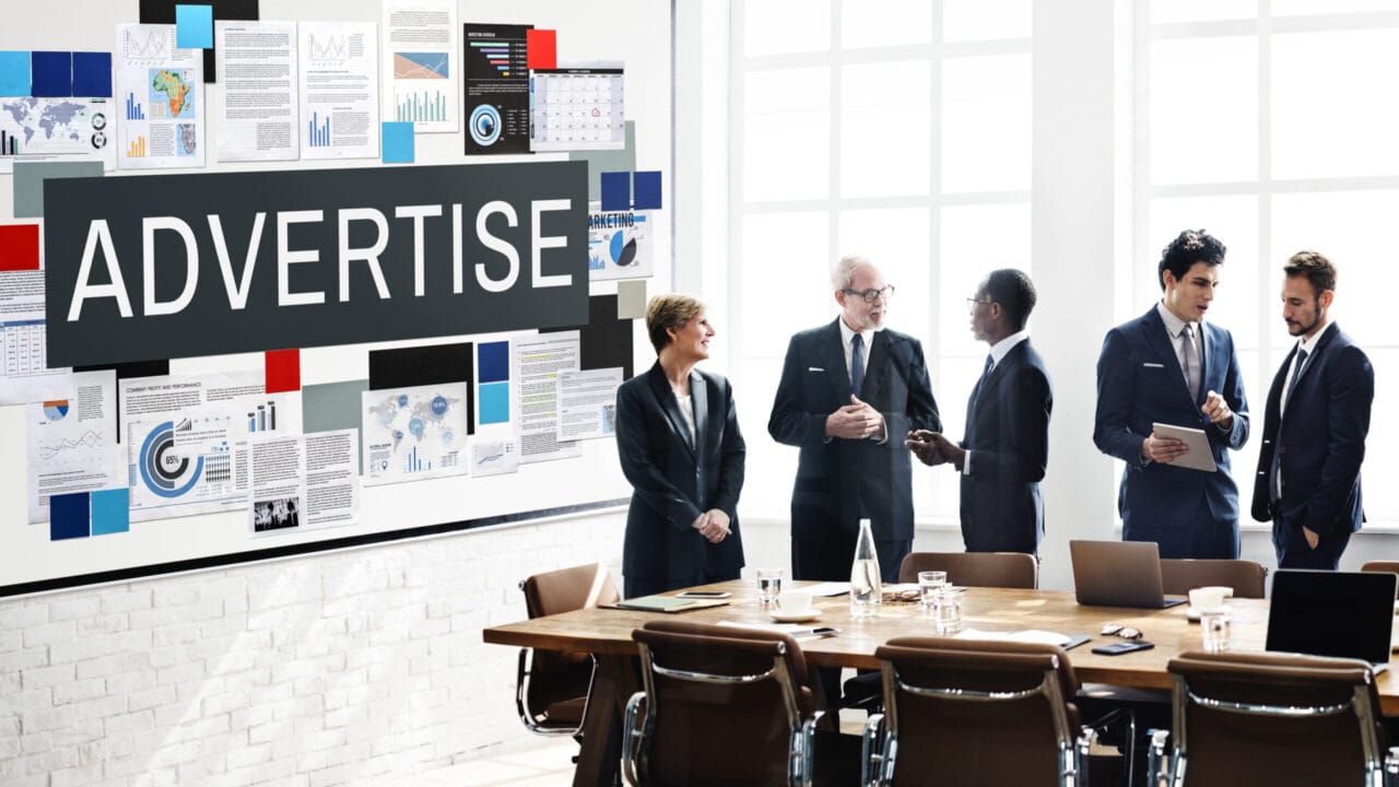 Team discussing marketing strategy in modern office with large "Advertise" sign, charts and laptop.