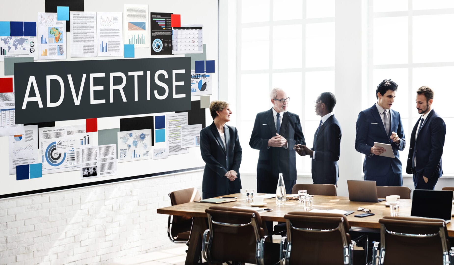 Team discussing marketing strategy in modern office with large "Advertise" sign, charts and laptop.