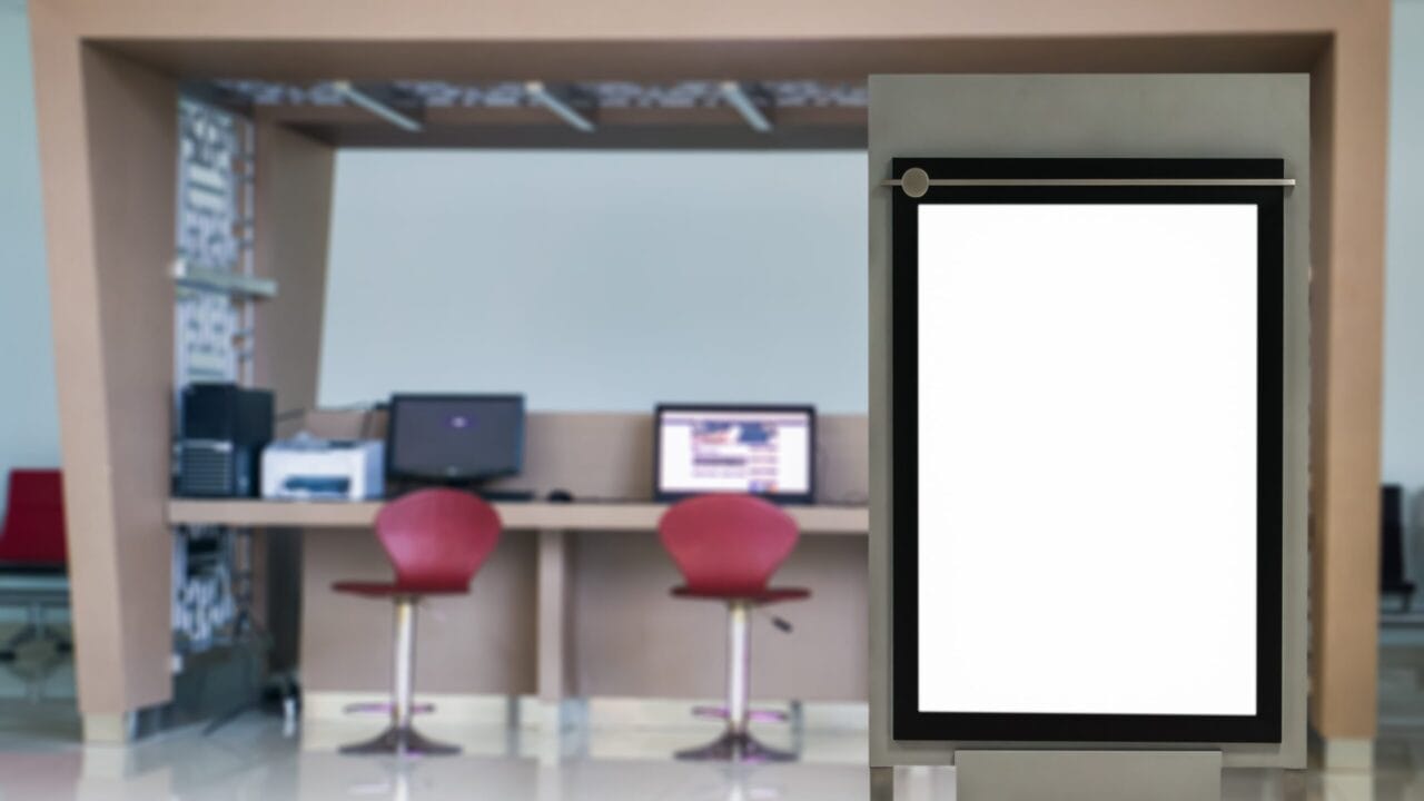 Blank digital kiosk sign in modern office lobby with desks and red chairs in the background.