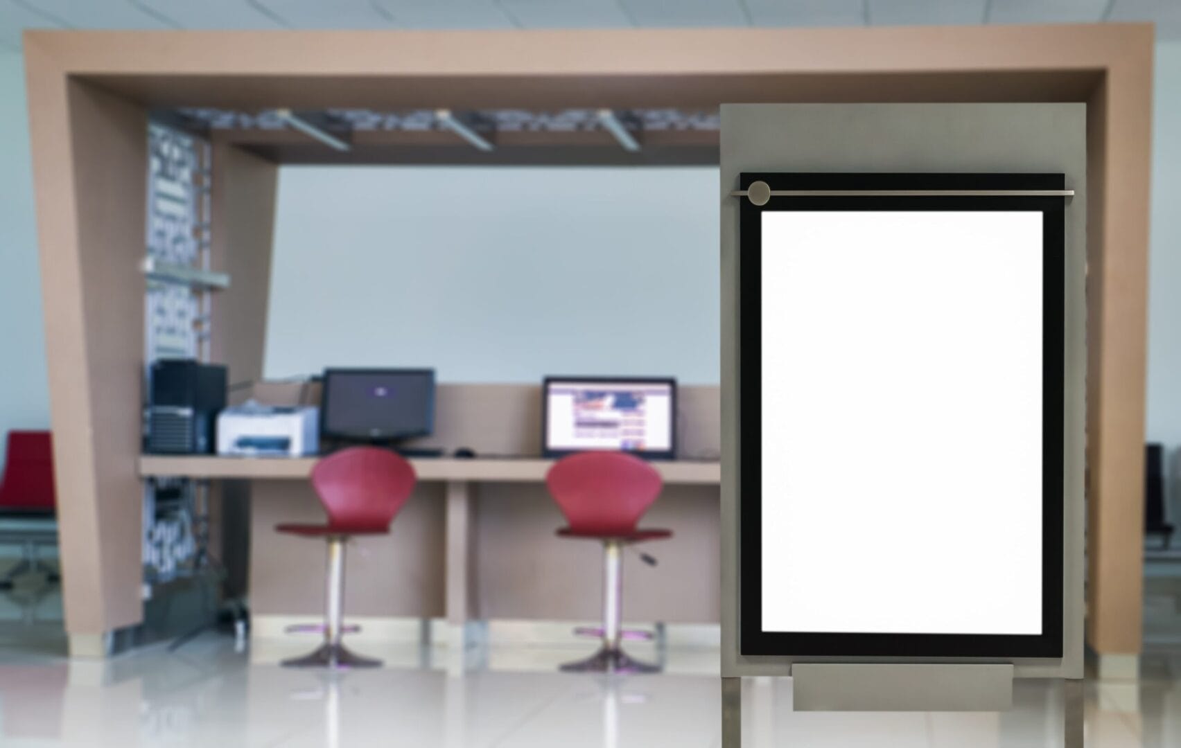 Blank digital kiosk sign in modern office lobby with desks and red chairs in the background.