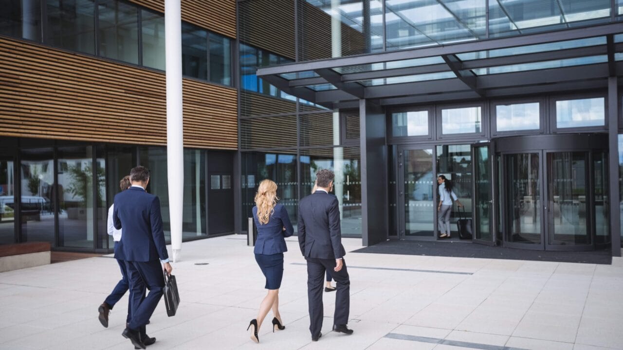Business professionals walking towards a modern office building entrance, carrying bags and wearing formal attire.
