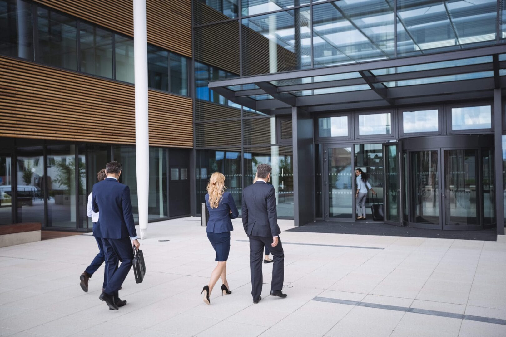 Business professionals walking towards a modern office building entrance, carrying bags and wearing formal attire.