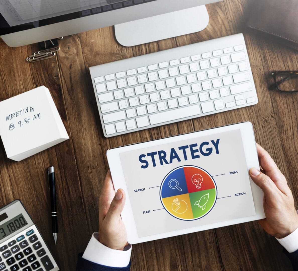 Person holding tablet displaying strategy diagram with colorful segments at a wooden desk with computer and meeting note.