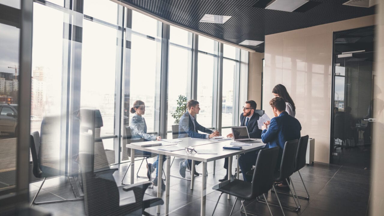 Business meeting in a modern office with a diverse team discussing strategy around a table, large windows in the background.