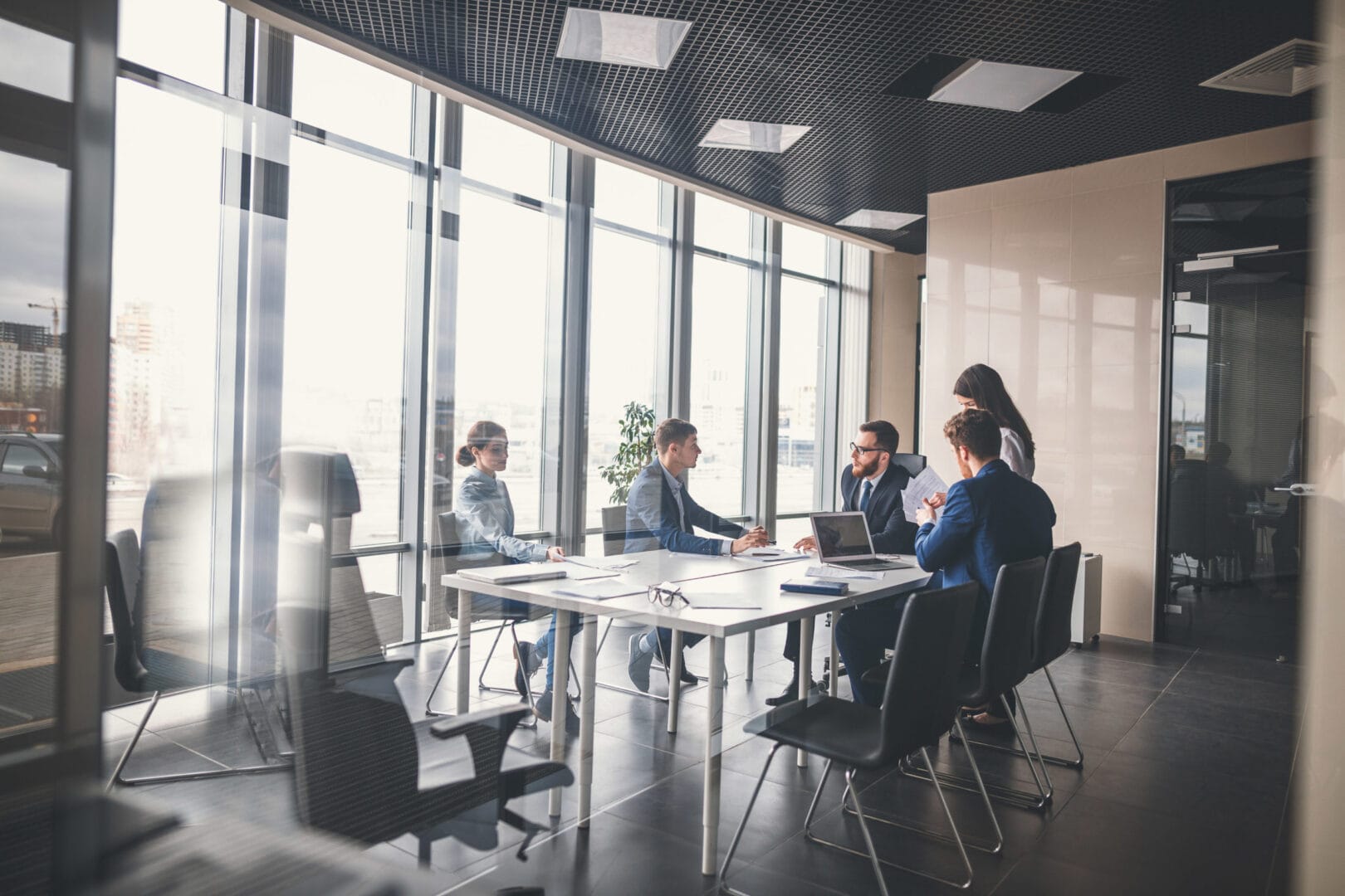 Business meeting in a modern office with a diverse team discussing strategy around a table, large windows in the background.