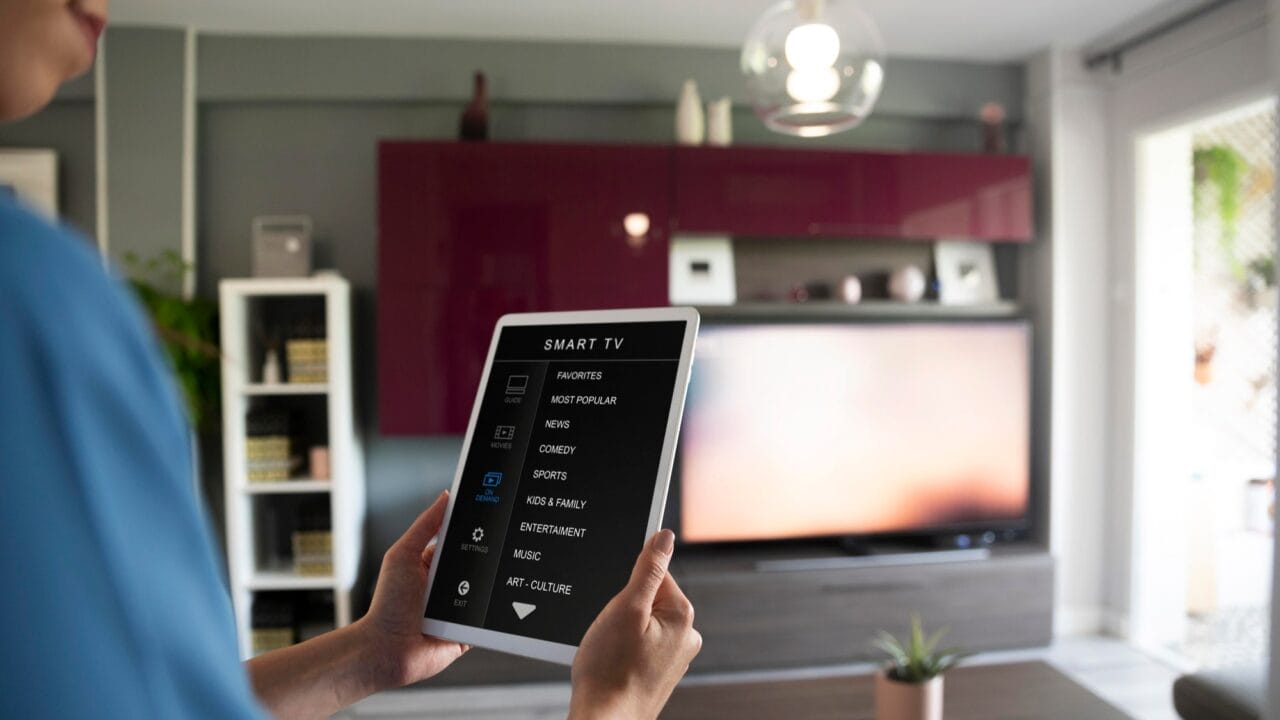 Person holding a tablet with smart TV controls in a modern living room with a flat-screen TV and contemporary decor.