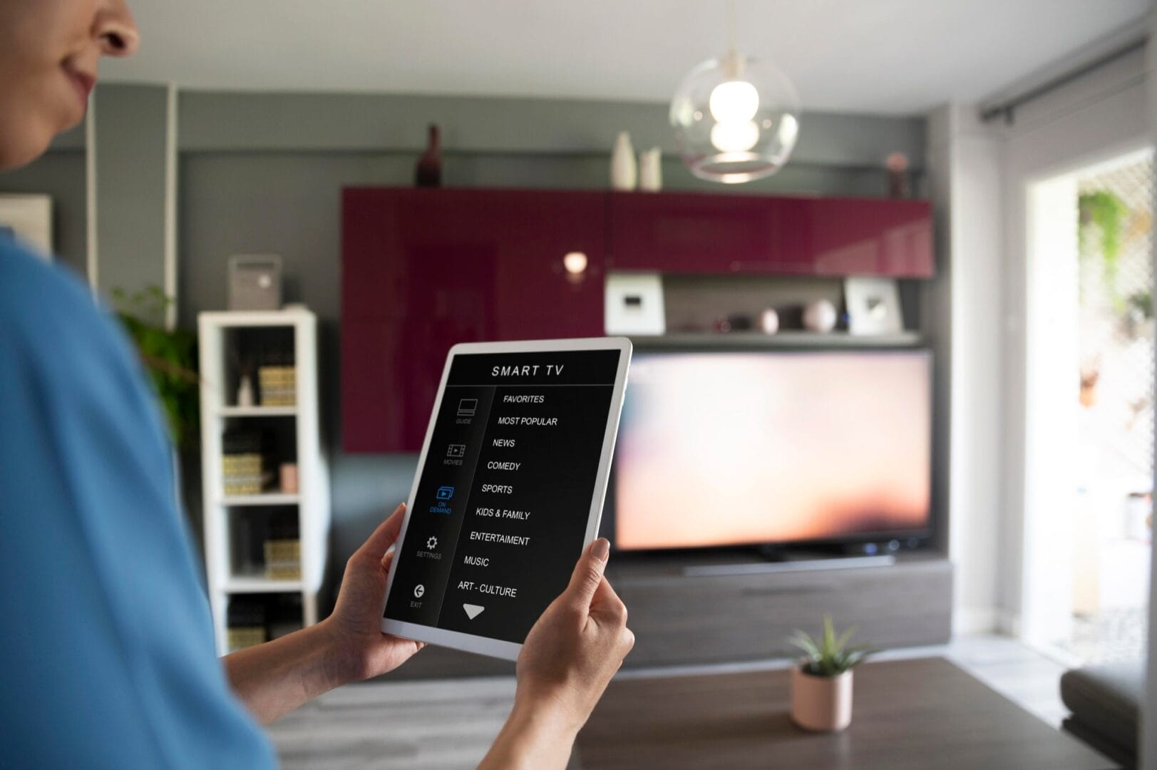 Person holding a tablet with smart TV controls in a modern living room with a flat-screen TV and contemporary decor.