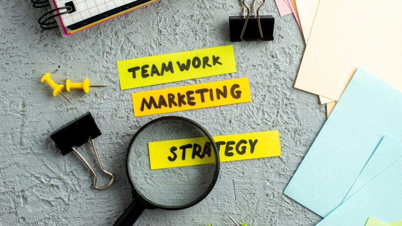 Magnifying glass over colorful notes reading teamwork, marketing, strategy on a textured gray background with office supplies.