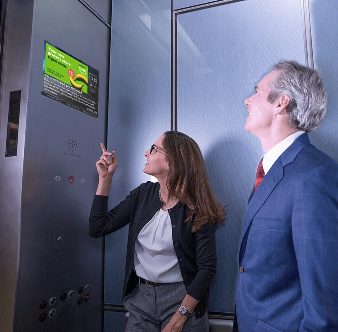 two people observing a digital screen inside an elevator displaying an advertisement, both dressed in business attire