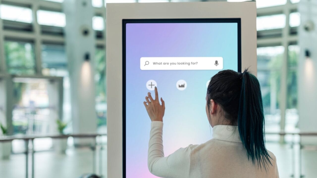 Person using a touchscreen kiosk with search bar in a modern building lobby.