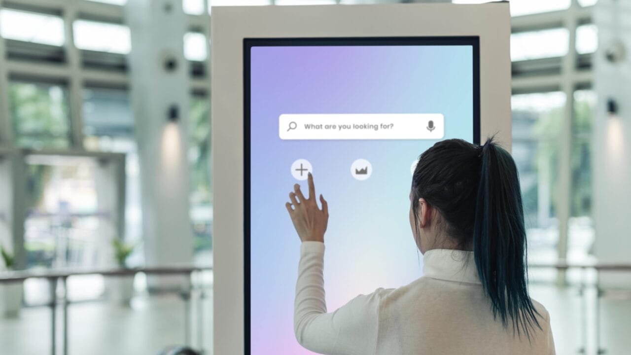 Person using a large touchscreen kiosk with a search bar interface in a modern building interior.