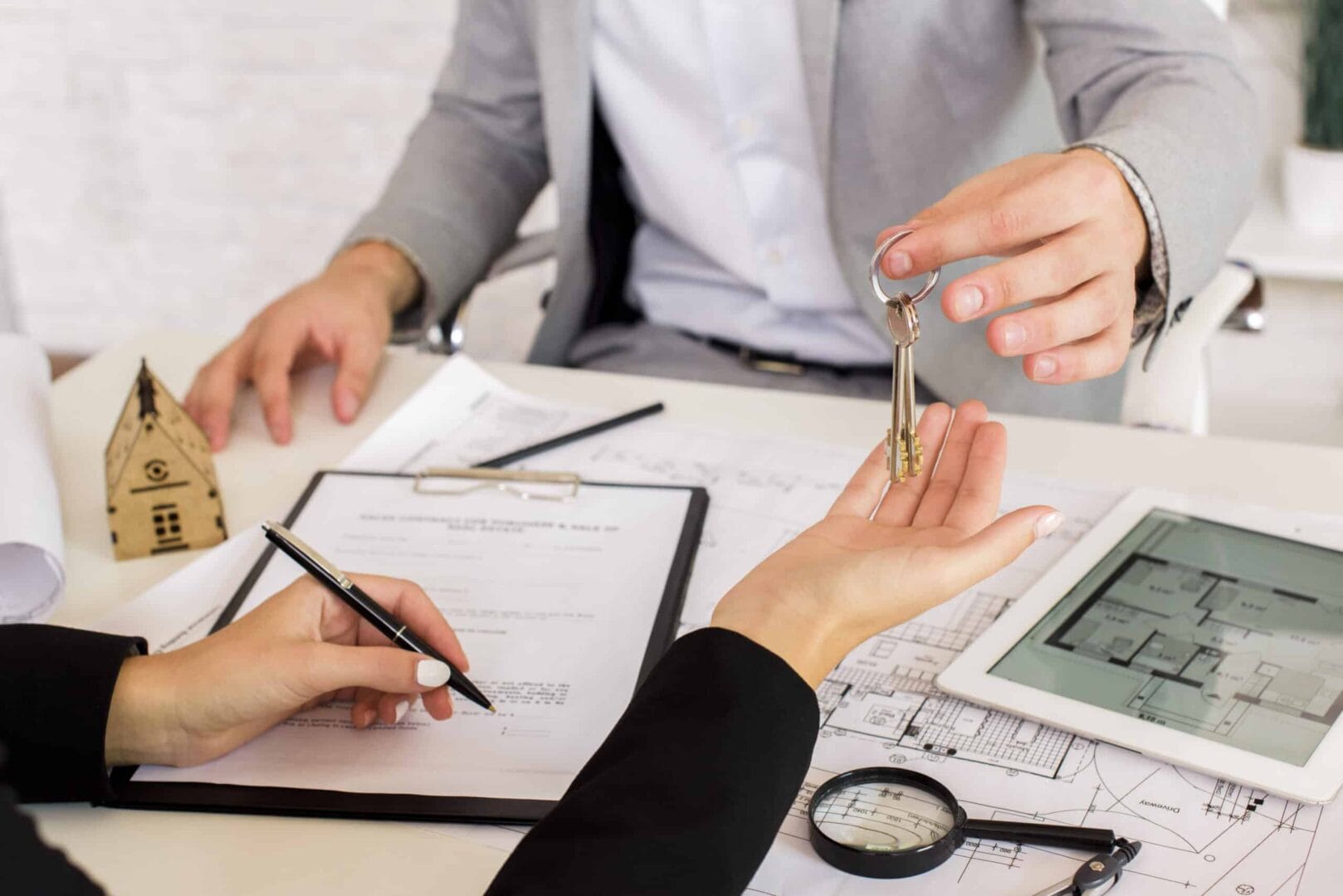 Real estate agent handing house keys to a new homeowner signing contract at a desk with house model and floor plans.