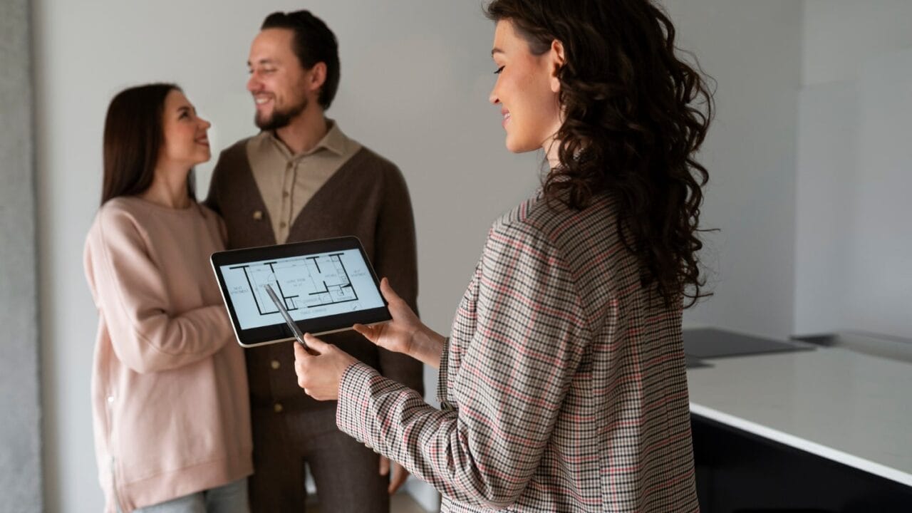 Real estate agent showing house plans on tablet to a smiling couple inside a home.