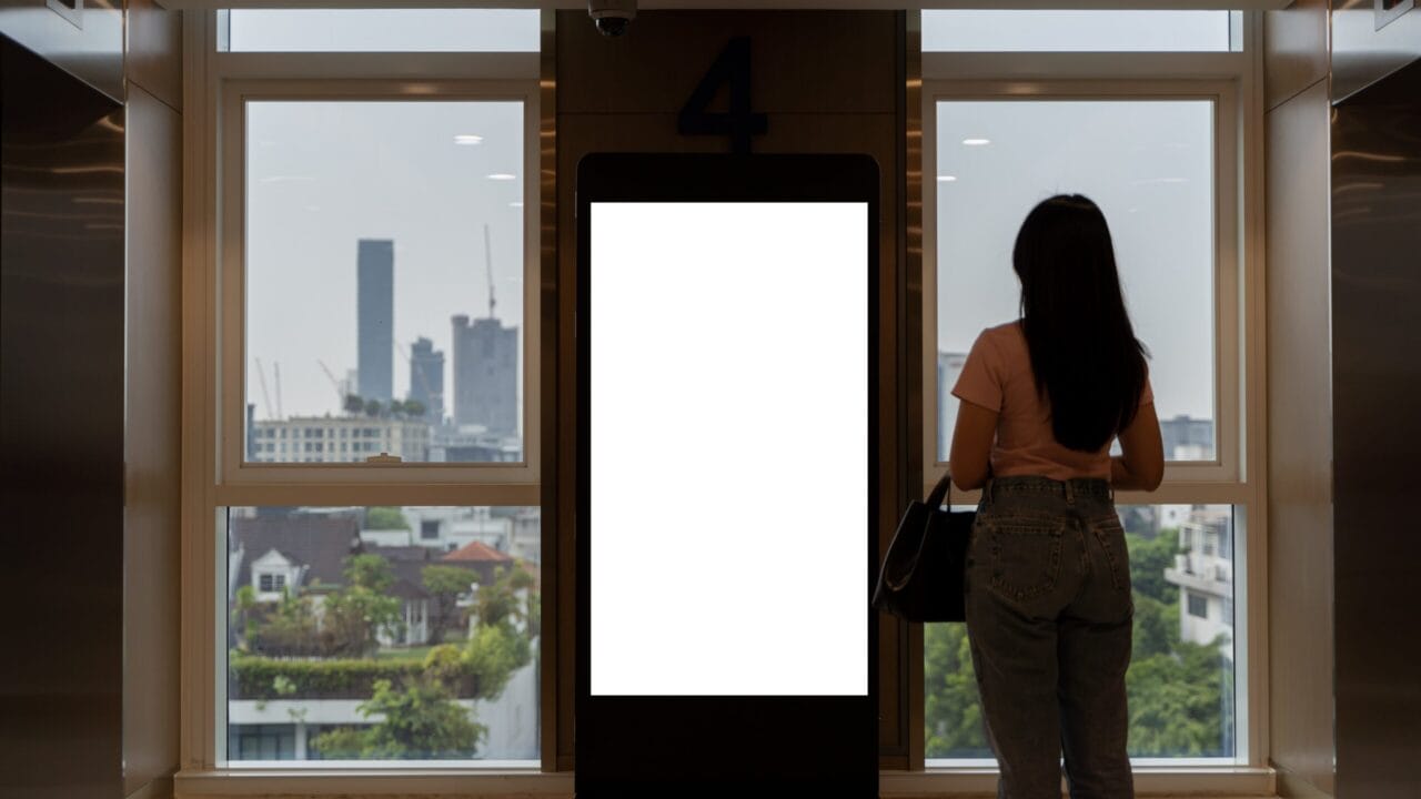 Woman standing near a blank digital billboard in a modern building lobby with a city view through large windows.