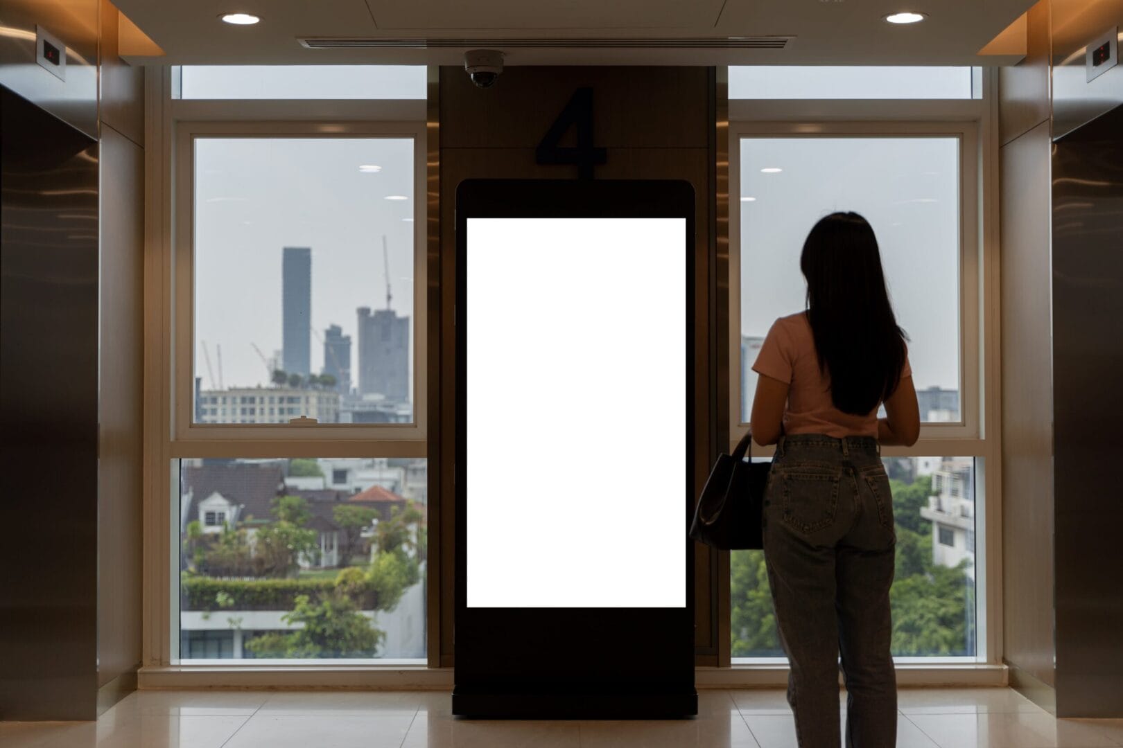 Woman standing near a blank digital billboard in a modern building lobby with a city view through large windows.