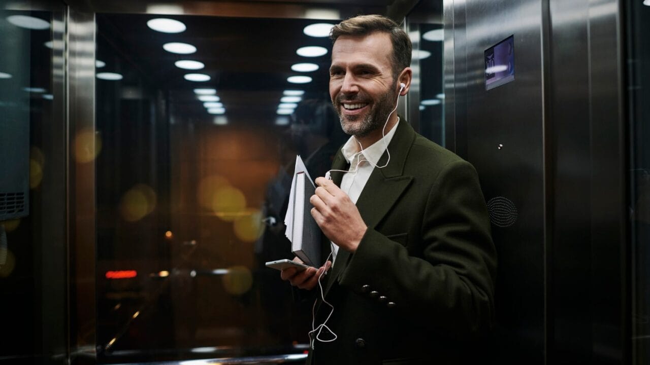 Man in suit smiling with headphones and holding a book in an elevator.